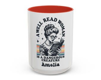 Può includere: Tazza in ceramica bianca con interno rosso. La tazza presenta un'illustrazione in bianco e nero di una donna che legge un libro, con il testo "A WELL READ WOMAN IS A DANGEROUS CREATURE Amelia" in un design circolare con una striscia arcobaleno retrò.