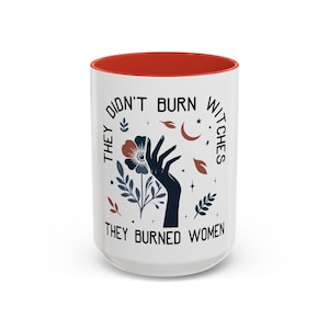 May include: A white ceramic mug with a red interior. The mug features the text "THEY DIDN'T BURN WITCHES THEY BURNED WOMEN" with a black hand, floral, and celestial design.