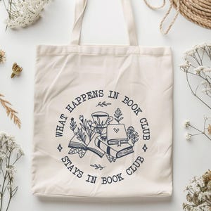 May include: A white canvas tote bag with a black and white graphic design. The graphic features a circle with the text "What happens in book club stays in book club" and an illustration of books, a coffee cup, and flowers.