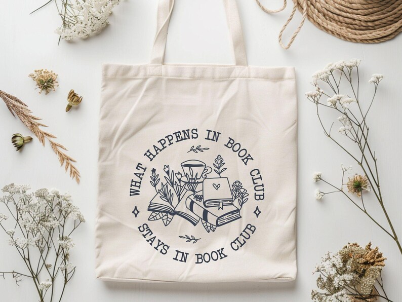 May include: A white canvas tote bag with a black and white graphic design. The graphic features a circle with the text "What happens in book club stays in book club" and an illustration of books, a coffee cup, and flowers.