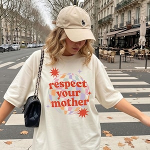 Könnte beinhalten: Eine beige Baseballkappe mit kleinem dunklem Logo und ein cremefarbenes T-Shirt mit einer Grafik einer karierten Discokugel und dem roten Schriftzug "respect your mother", umgeben von Sternformen, kombiniert mit einer schwarzen gesteppten Umhängetasche.