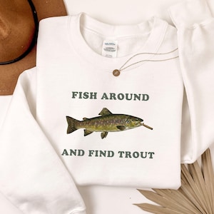 May include: White sweatshirt featuring the text "FISH AROUND AND FIND TROUT" in dark green. A detailed illustration of a trout with a cigarette in its mouth is centered below the text. A brown hat and necklace are visible.