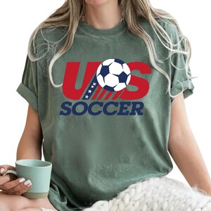 Puede incluir: Una camiseta de manga corta color verde salvia con un dise&ntilde;o gr&aacute;fico que presenta el texto rojo US, un bal&oacute;n de f&uacute;tbol azul y blanco que reemplaza la letra O, y la palabra SOCCER impresa en azul marino debajo. El dise&ntilde;o incluye cuatro estrellas blancas.