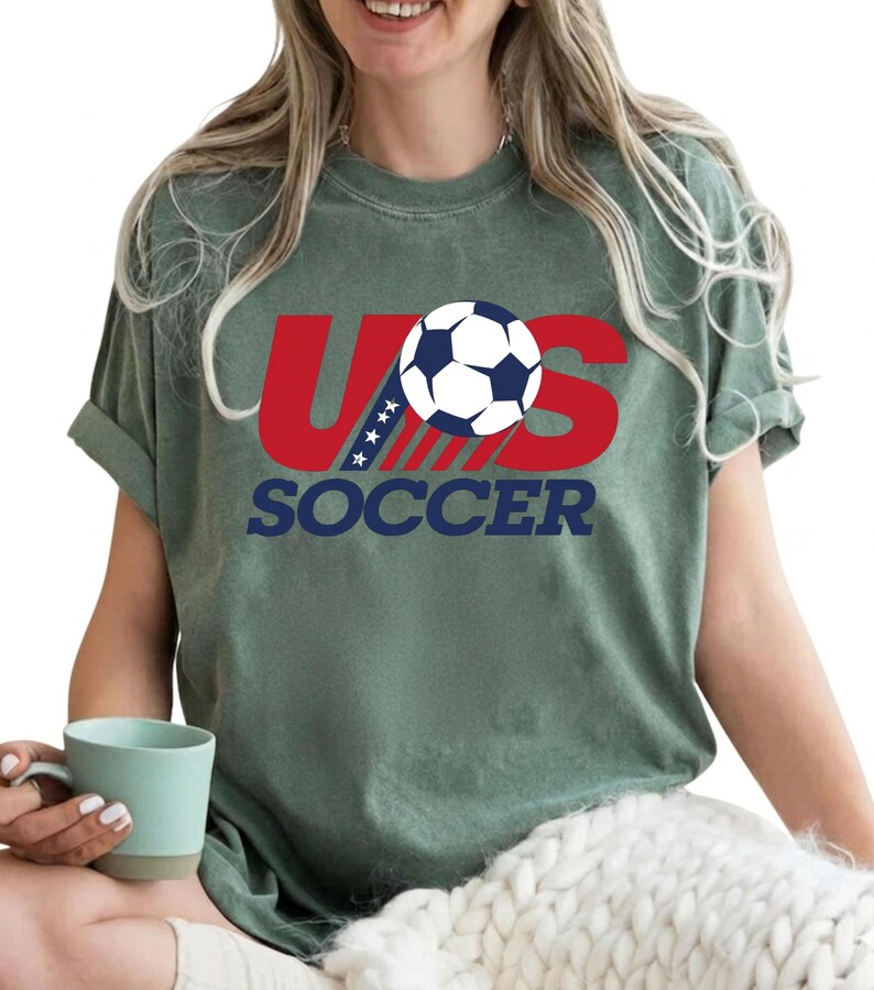 Puede incluir: Una camiseta de manga corta color verde salvia con un dise&ntilde;o gr&aacute;fico que presenta el texto rojo US, un bal&oacute;n de f&uacute;tbol azul y blanco que reemplaza la letra O, y la palabra SOCCER impresa en azul marino debajo. El dise&ntilde;o incluye cuatro estrellas blancas.