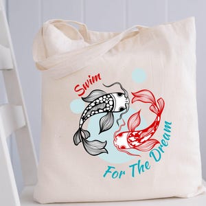 May include: Cream-colored tote bag featuring a black and red koi fish design with the text "Swim For The Dream". The fish are arranged in a circular pattern on a light blue background. The bag has two handles.