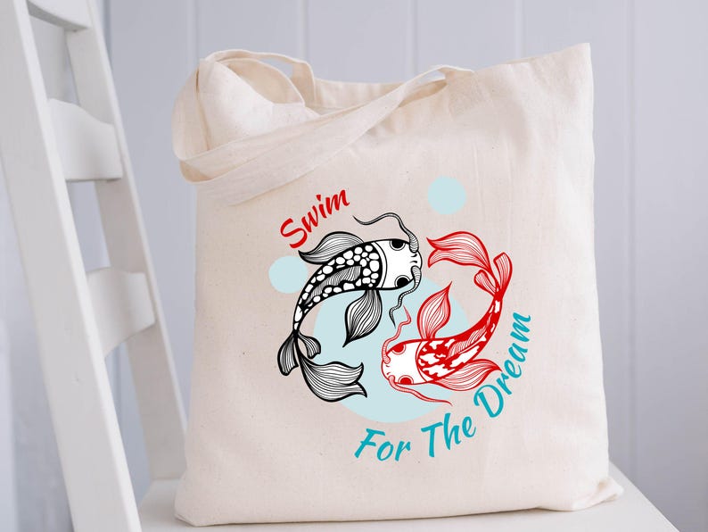 May include: Cream-colored tote bag featuring a black and red koi fish design with the text "Swim For The Dream". The fish are arranged in a circular pattern on a light blue background. The bag has two handles.
