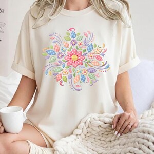 Könnte beinhalten: Cremefarbenes T-Shirt mit einem farbenfrohen Blumendesign. Das Design umfasst eine große rosa Blume mit gelbem Zentrum, umgeben von Blättern und floralen Elementen in Pastellfarben. Das Shirt hat kurze Ärmel und eine lockere Passform.