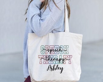 May include: A white canvas tote bag with the text "SPEECH THERAPY" in rainbow colors and the name "Ashley" in black script.