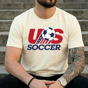Puede incluir: Una camiseta de manga corta de color crema con un dise&ntilde;o gr&aacute;fico que presenta el texto rojo US, un bal&oacute;n de f&uacute;tbol azul y blanco que sustituye a la letra O, y la palabra SOCCER en azul debajo. El dise&ntilde;o incluye estrellas y rayas azules y blancas.