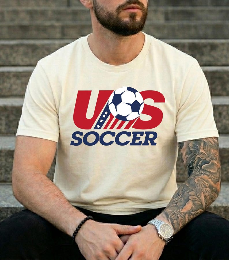 Puede incluir: Una camiseta de manga corta de color crema con un dise&ntilde;o gr&aacute;fico que presenta el texto rojo US, un bal&oacute;n de f&uacute;tbol azul y blanco que sustituye a la letra O, y la palabra SOCCER en azul debajo. El dise&ntilde;o incluye estrellas y rayas azules y blancas.