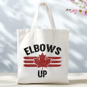 Puede incluir: Bolsa de lona blanca con texto negro que dice "Elbows Up" y un gráfico de hoja de arce roja.