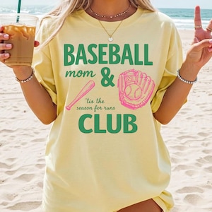 Op de afbeelding: Een lichtgeel t-shirt met de woorden "BASEBALL mom & CLUB" in het groen, en "'tis the season for runs" in het groen. Een roze honkbalknuppel en handschoen staan ook op het shirt.