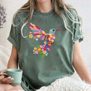 May include: Olive green t-shirt with a vibrant hummingbird design. The bird is made up of patchwork patterns in multiple colours, with floral details. The t-shirt has short sleeves and a relaxed fit, suitable for casual wear.