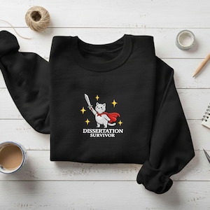 May include: Black sweatshirt featuring a cartoon cat in a red cape holding a sword, with the text "Dissertation Survivor" below. The design includes yellow star accents. The sweatshirt is displayed on a white wooden surface.