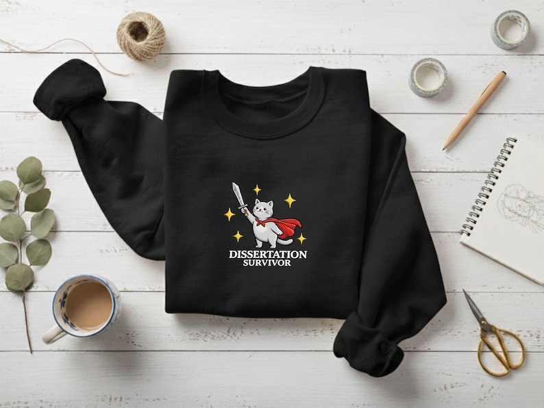 May include: Black sweatshirt featuring a cartoon cat in a red cape holding a sword, with the text "Dissertation Survivor" below. The design includes yellow star accents. The sweatshirt is displayed on a white wooden surface.