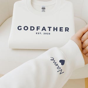 Pu&ograve; includere: Una felpa bianca piegata con la scritta "GODFATHER" e l'anno "EST. 2025" stampati in blu navy. La manica di un'altra felpa bianca mostra il nome "Name" e un cuore blu navy.