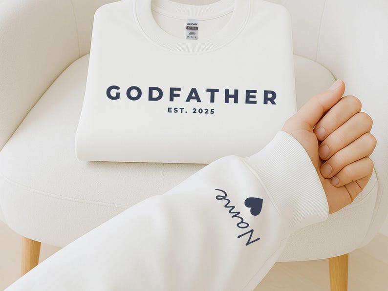 Pu&ograve; includere: Una felpa bianca piegata con la scritta "GODFATHER" e l'anno "EST. 2025" stampati in blu navy. La manica di un'altra felpa bianca mostra il nome "Name" e un cuore blu navy.