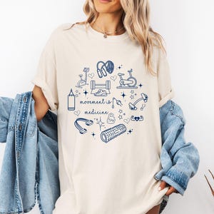 Può includere: T-shirt color crema con grafica blu navy di attrezzature per il fitness e la scritta "movement is medicine". Il design include un sacco da boxe, cyclette, manubri e un rullo in schiuma. La maglietta è in stile casual e confortevole.