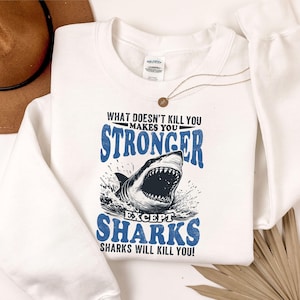 May include: White sweatshirt with a shark graphic and the text "WHAT DOESN'T KILL YOU MAKES YOU STRONGER EXCEPT SHARKS SHARKS WILL KILL YOU!". A brown hat and necklace are also visible.