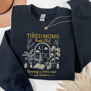 Op de afbeelding: Zwart sweatshirt met de gouden tekst "Tired Moms Book Club". Het ontwerp bevat een raam met een maan, boeken, planten en kaarsen. De zin "Running on Coffee, Chaos and chapters" staat er ook op.