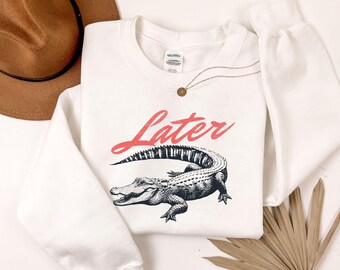 May include: White sweatshirt with the word "Later" in red script above a detailed black and white alligator illustration. A gold necklace with a small pendant is draped on the shirt.