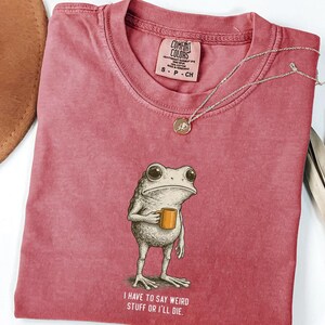 May include: A dusty rose Comfort Colors t-shirt with a frog illustration holding a mug. The frog has large eyes and is standing upright. The text "I HAVE TO SAY WEIRD STUFF OR I'LL DIE" is printed below the frog. A silver necklace with a pendant is on the shirt.