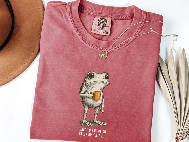 May include: A dusty rose Comfort Colors t-shirt with a frog illustration holding a mug. The frog has large eyes and is standing upright. The text "I HAVE TO SAY WEIRD STUFF OR I'LL DIE" is printed below the frog. A silver necklace with a pendant is on the shirt.