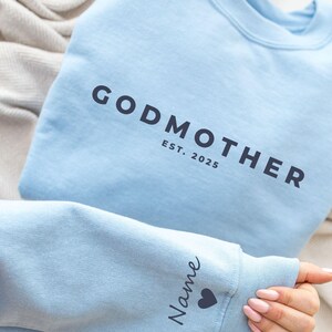 Pu&ograve; includere: Felpa azzurra con la scritta "GODMOTHER EST. 2025" stampata in grigio scuro. La manica presenta la parola "Name" e un simbolo a forma di cuore. La felpa &egrave; piegata su una superficie colorata neutra.
