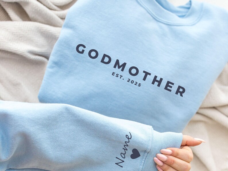 Pu&ograve; includere: Felpa azzurra con la scritta "GODMOTHER EST. 2025" stampata in grigio scuro. La manica presenta la parola "Name" e un simbolo a forma di cuore. La felpa &egrave; piegata su una superficie colorata neutra.