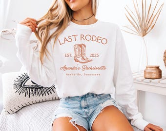 May include: White long-sleeved sweatshirt featuring a brown graphic with the words "LAST RODEO", a pair of cowboy boots, and the text "Amanda's Bachelorette" and "Nashville, Tennessee". The year "2025" is also printed on the shirt.