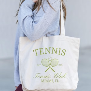 Könnte beinhalten: Cremefarbene Tragetasche mit grünem Text und Grafiken. Das Wort "TENNIS" steht oben, mit gekreuzten Tennisschlägern und einem Ball darunter. Der Text "Tennis Club MIAMI, FL" steht unten, mit "EST. 2025" auf beiden Seiten der Schläger.