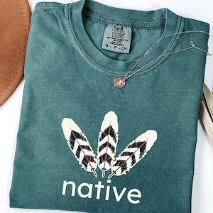 Op de afbeelding: Tealkleurig T-shirt met een afbeelding van drie veren en het woord "native" in het wit. Een gouden ketting met een hanger ligt op het shirt. Een bruine hoed is zichtbaar op de achtergrond.