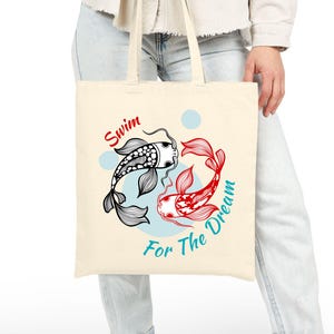 May include: Cream-colored tote bag featuring a black and white koi fish and a red koi fish in a circular design. The words "Swim" and "For The Dream" are written in red and teal, respectively.