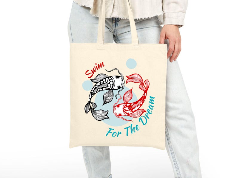 May include: Cream-colored tote bag featuring a black and white koi fish and a red koi fish in a circular design. The words "Swim" and "For The Dream" are written in red and teal, respectively.