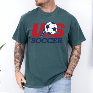 Puede incluir: Una camiseta de manga corta color verde azulado oscuro con un dise&ntilde;o gr&aacute;fico que muestra las letras rojas US, un bal&oacute;n de f&uacute;tbol blanco y azul que sustituye a la letra O, y la palabra SOCCER en azul oscuro debajo. Incluye cuatro estrellas blancas.