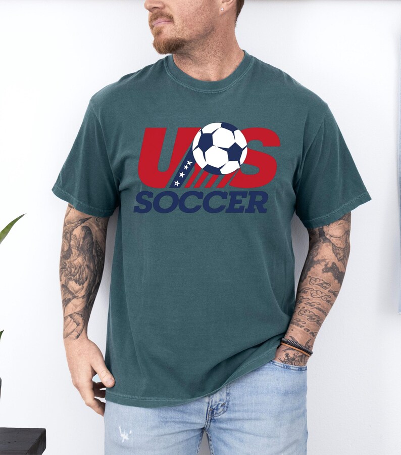 Puede incluir: Una camiseta de manga corta color verde azulado oscuro con un dise&ntilde;o gr&aacute;fico que muestra las letras rojas US, un bal&oacute;n de f&uacute;tbol blanco y azul que sustituye a la letra O, y la palabra SOCCER en azul oscuro debajo. Incluye cuatro estrellas blancas.