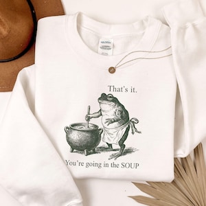 May include: White sweatshirt with a frog illustration stirring a pot, with the text "That's it. You're going in the SOUP." A gold necklace and a brown hat are also in the image. The sweatshirt has long sleeves.