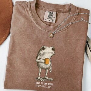 May include: Brown Comfort Colors t-shirt featuring a frog graphic holding a mug, with the text "I have to say weird stuff or I'll die". A silver necklace and a hat are also visible in the image.