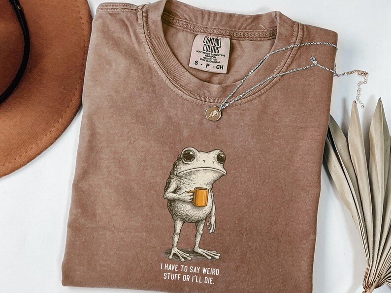 May include: Brown Comfort Colors t-shirt featuring a frog graphic holding a mug, with the text "I have to say weird stuff or I'll die". A silver necklace and a hat are also visible in the image.