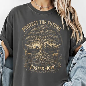 May include: Dark gray t-shirt with a nature-themed graphic. The design features a tree with a mountain backdrop, deer, and the text "PROTECT THE FUTURE" and "FOSTER HOPE" in a circular layout. The shirt has a relaxed fit.
