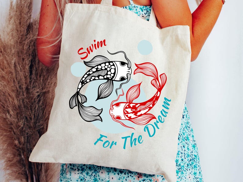 May include: Cream-colored tote bag featuring a graphic of two koi fish in black and red, with the words "Swim For The Dream" in red and teal. The design includes two blue circles.