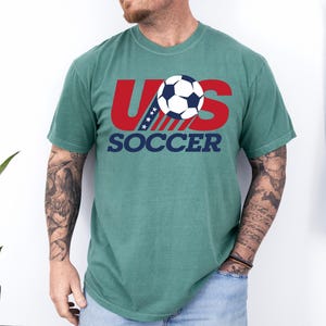 Puede incluir: Una camiseta de manga corta color verde salvia con un gr&aacute;fico en el pecho. El gr&aacute;fico muestra el texto rojo US con estrellas y rayas azules, un bal&oacute;n de f&uacute;tbol blanco y negro, y la palabra SOCCER en letras azul marino debajo del dise&ntilde;o.