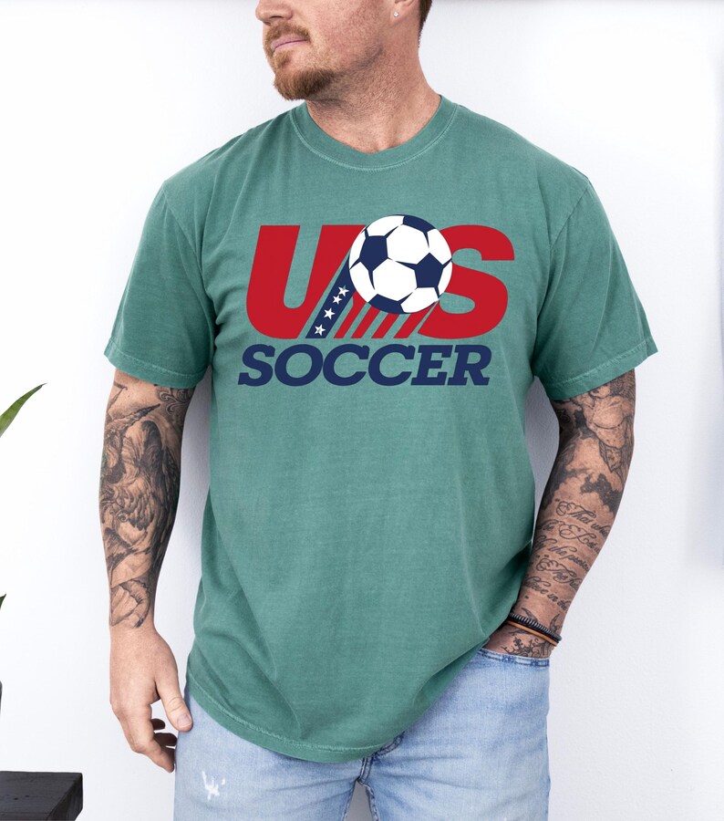 Puede incluir: Una camiseta de manga corta color verde salvia con un gr&aacute;fico en el pecho. El gr&aacute;fico muestra el texto rojo US con estrellas y rayas azules, un bal&oacute;n de f&uacute;tbol blanco y negro, y la palabra SOCCER en letras azul marino debajo del dise&ntilde;o.
