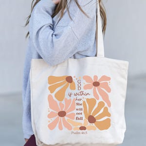 May include: A white canvas tote bag with a floral design and the text "GOD is within her. She will not fall. Psalm 46:5".