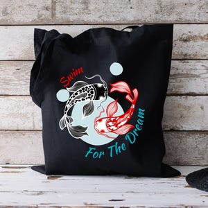 May include: Black tote bag featuring a koi fish design. One fish is black and white, the other is red and white. The text "Swim For The Dream" is printed on the bag in red and teal.