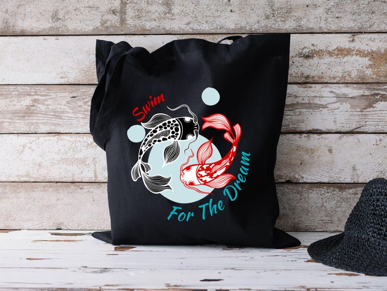 May include: Black tote bag featuring a koi fish design. One fish is black and white, the other is red and white. The text "Swim For The Dream" is printed on the bag in red and teal.