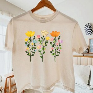May include: Cream-coloured t-shirt with a floral design. The design features three stems with colourful, patchwork-style flowers in yellow, pink, and orange. The flowers have green stems and leaves. The shirt is hanging on a wooden hanger.