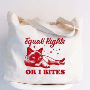 Peut inclure: Un sac fourre-tout en toile blanche avec du texte rouge qui dit "Equal Rights or I Bites". Une image de dessin animé d'un chat siamois aux yeux rouges est allongé sur le sac.
