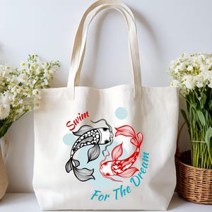 May include: Cream-colored tote bag featuring a graphic of two koi fish in black and red, with the words "Swim For The Dream" in red and teal. The bag has long handles and is displayed with white flowers.