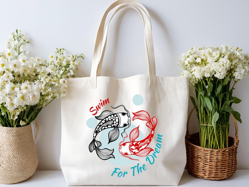 May include: Cream-colored tote bag featuring a graphic of two koi fish in black and red, with the words "Swim For The Dream" in red and teal. The bag has long handles and is displayed with white flowers.